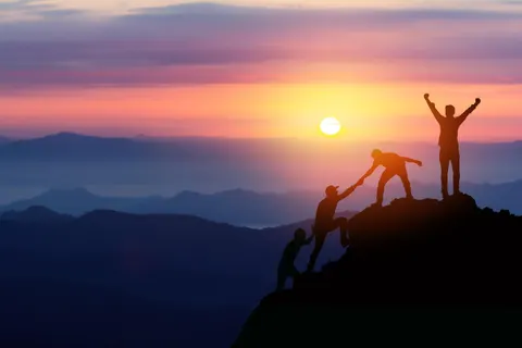 Silhouettes of climbers helping each other reach a mountain summit at sunrise